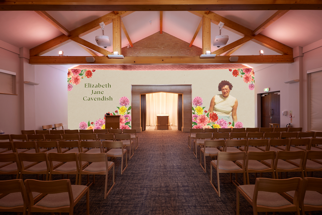 An indoor event space with wooden beams on the ceiling, rows of chairs facing a large screen displaying the name 'Elizabeth Jane Cavendish' surrounded by colorful pink, red, and yellow flowers, and a woman wearing a sleeveless beige top standing on the right side of the screen.