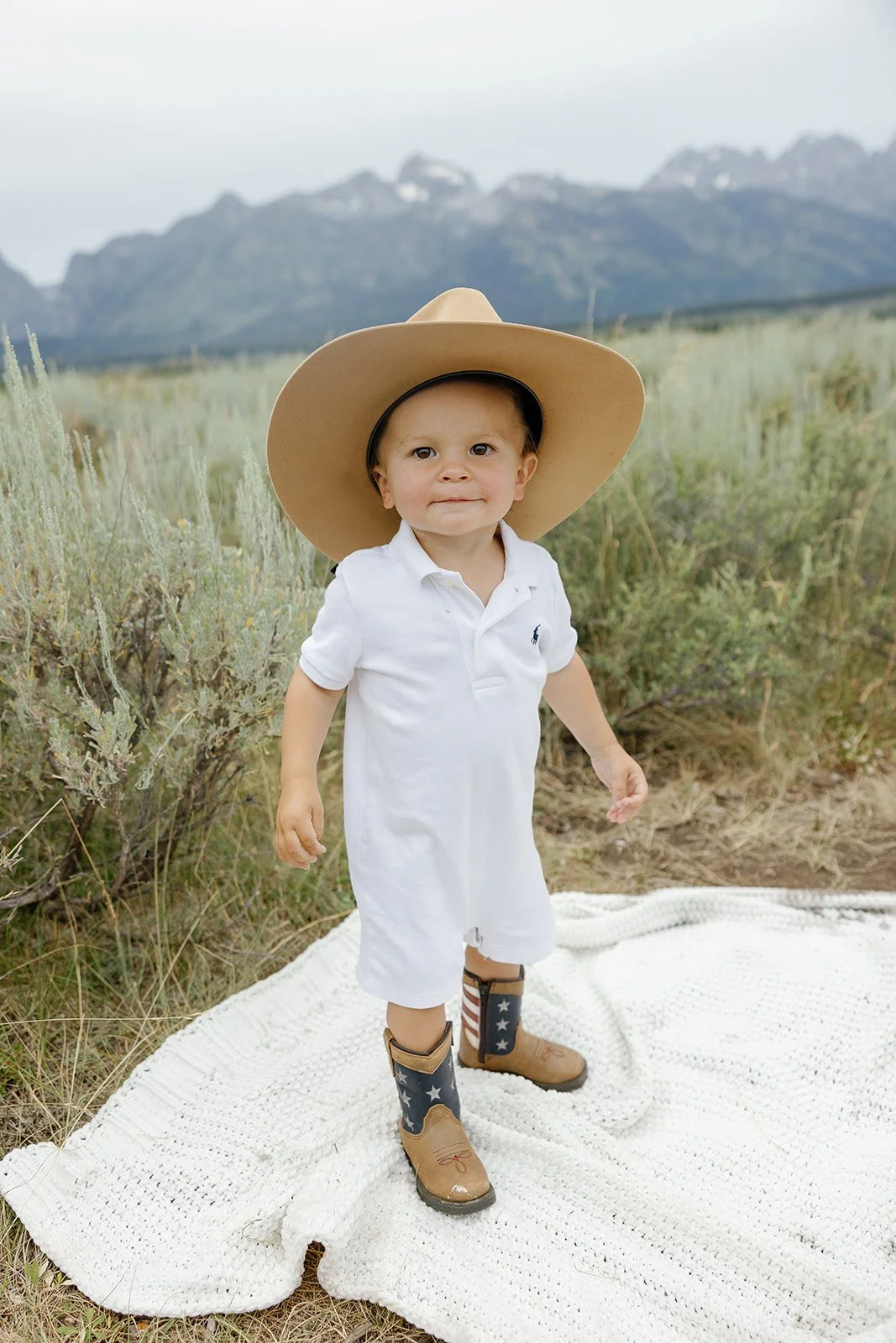 A young boy wearing a large floppy hat, a white jumpsuit, cowboy boots with an American flag design, standing on a white blanket in a natural outdoor setting with mountains in the background.