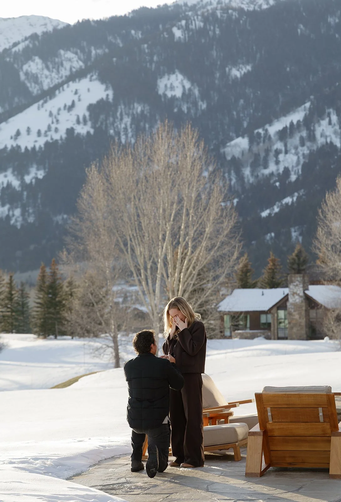 A man kneeling proposes to a woman outdoors in a snowy setting with mountain and house in the background.