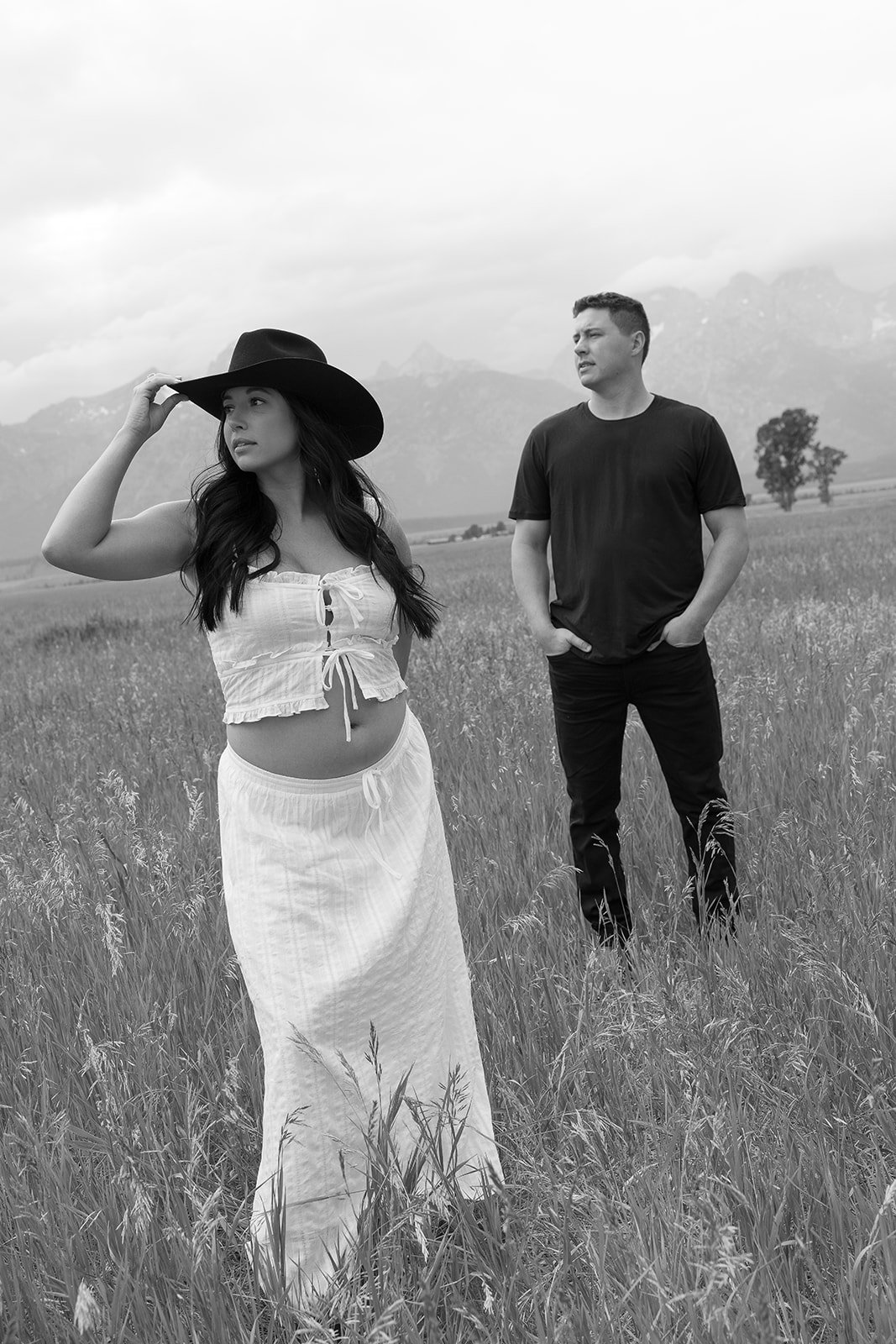 A woman in a large black hat, white crop top, and long skirt stands in a grassy field, with a man in a black shirt and pants standing behind her. Mountains are visible in the background. The image is in black and white.
