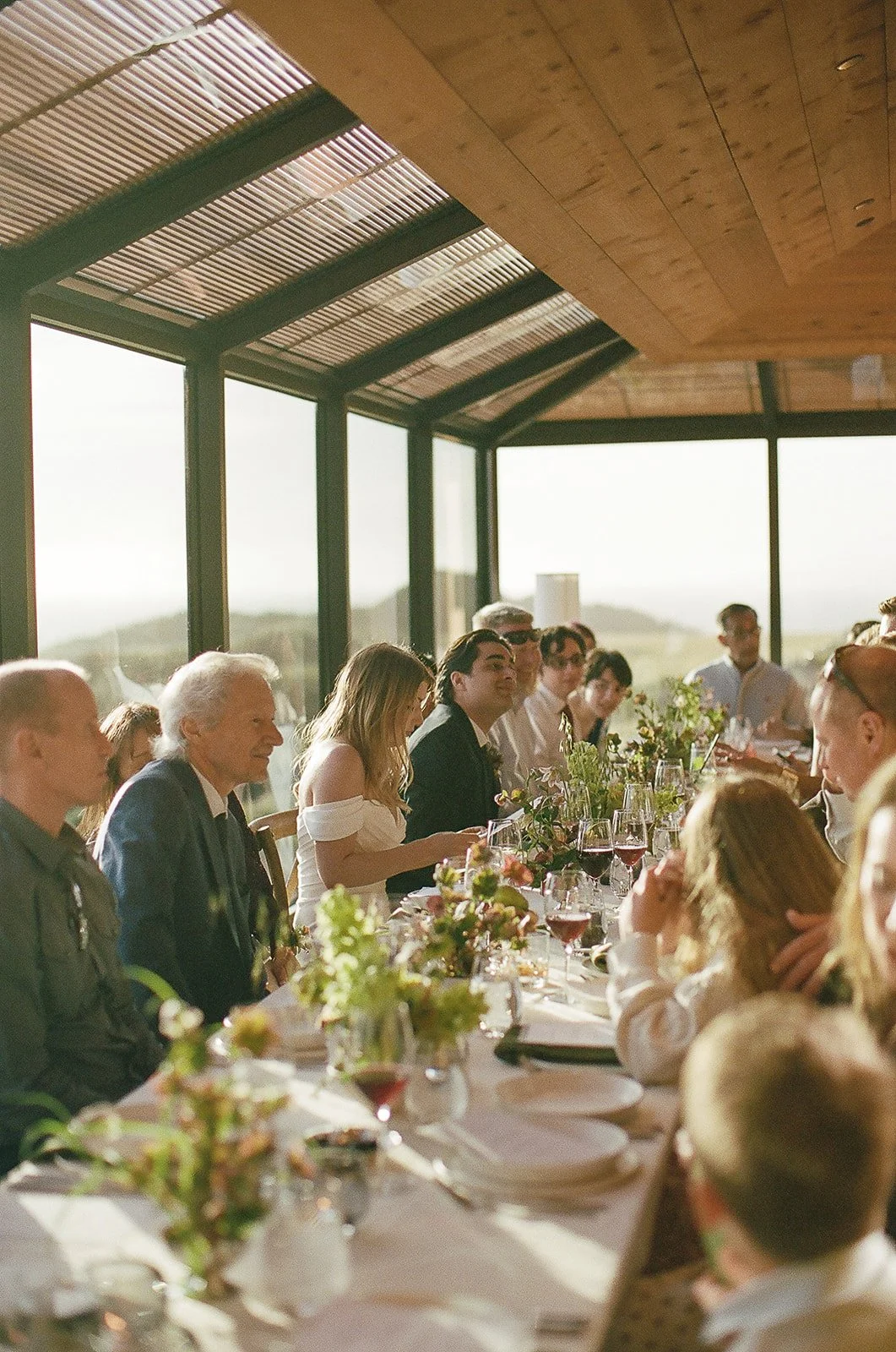 People sitting at a long table during a meal in a glass-walled room with a wooden ceiling, sunlight streaming in.