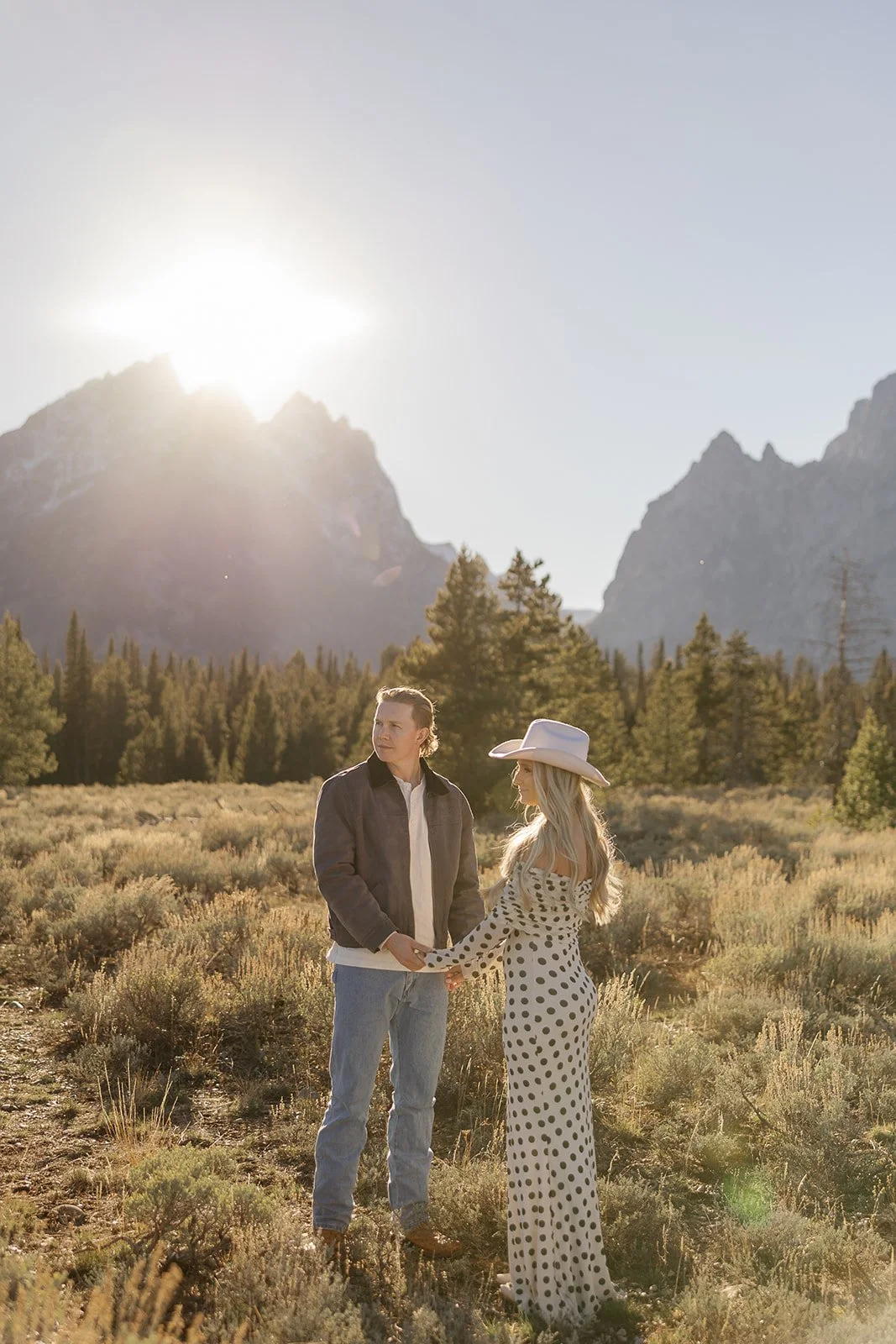 A man and woman holding hands in a grassy field with mountains and trees in the background, under a bright sun.