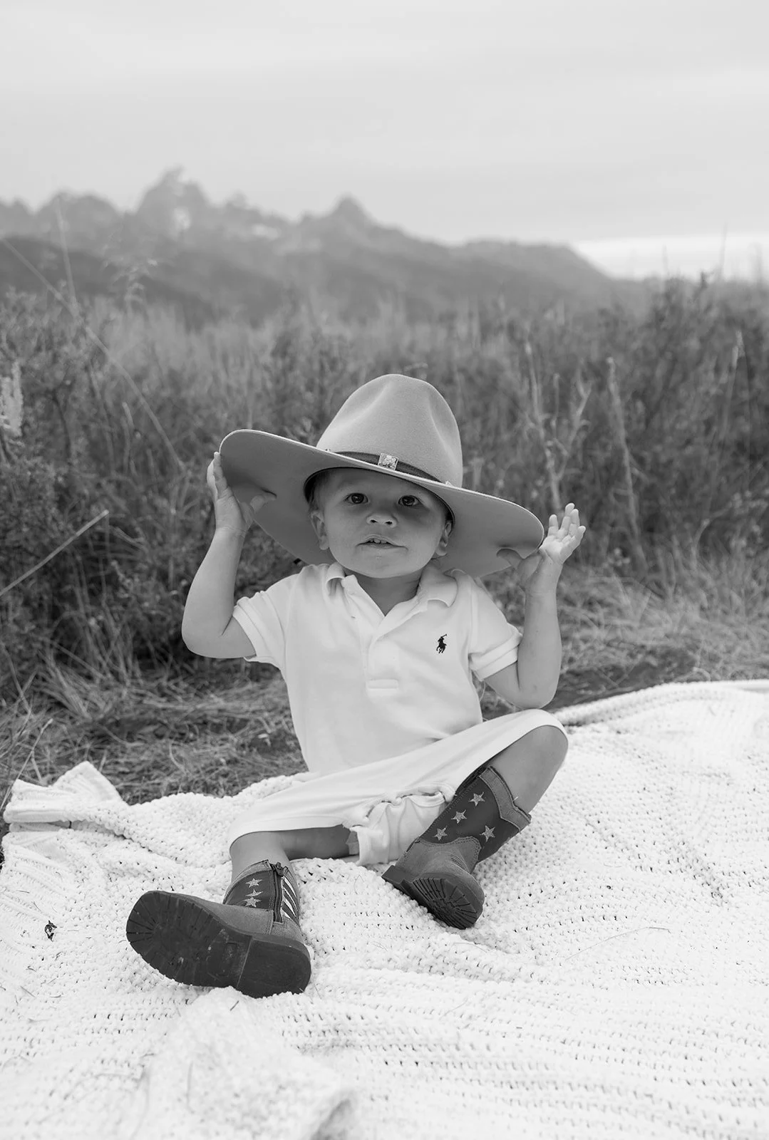 A young child sitting on a blanket outdoors, wearing cowboy boots and a large cowboy hat, with tall grass and a mountain in the background.