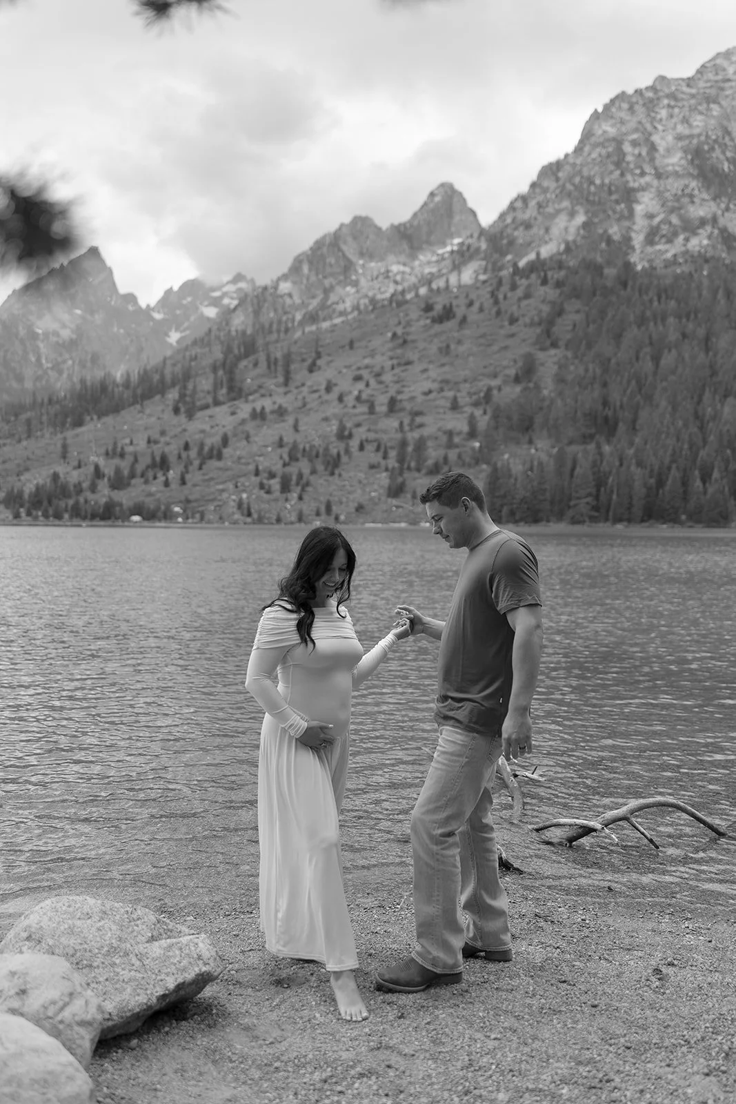 A couple standing by a lake, with mountains and trees in the background, with cloudy skies above.