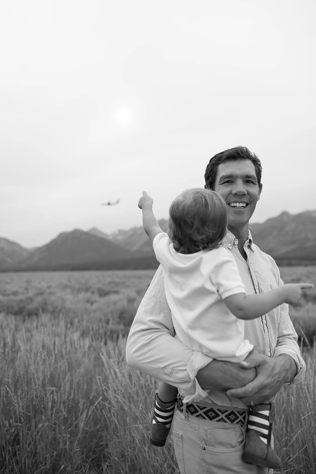 A man smiling while holding a young child in a grassy field with mountains in the background. An airplane is flying in the sky.