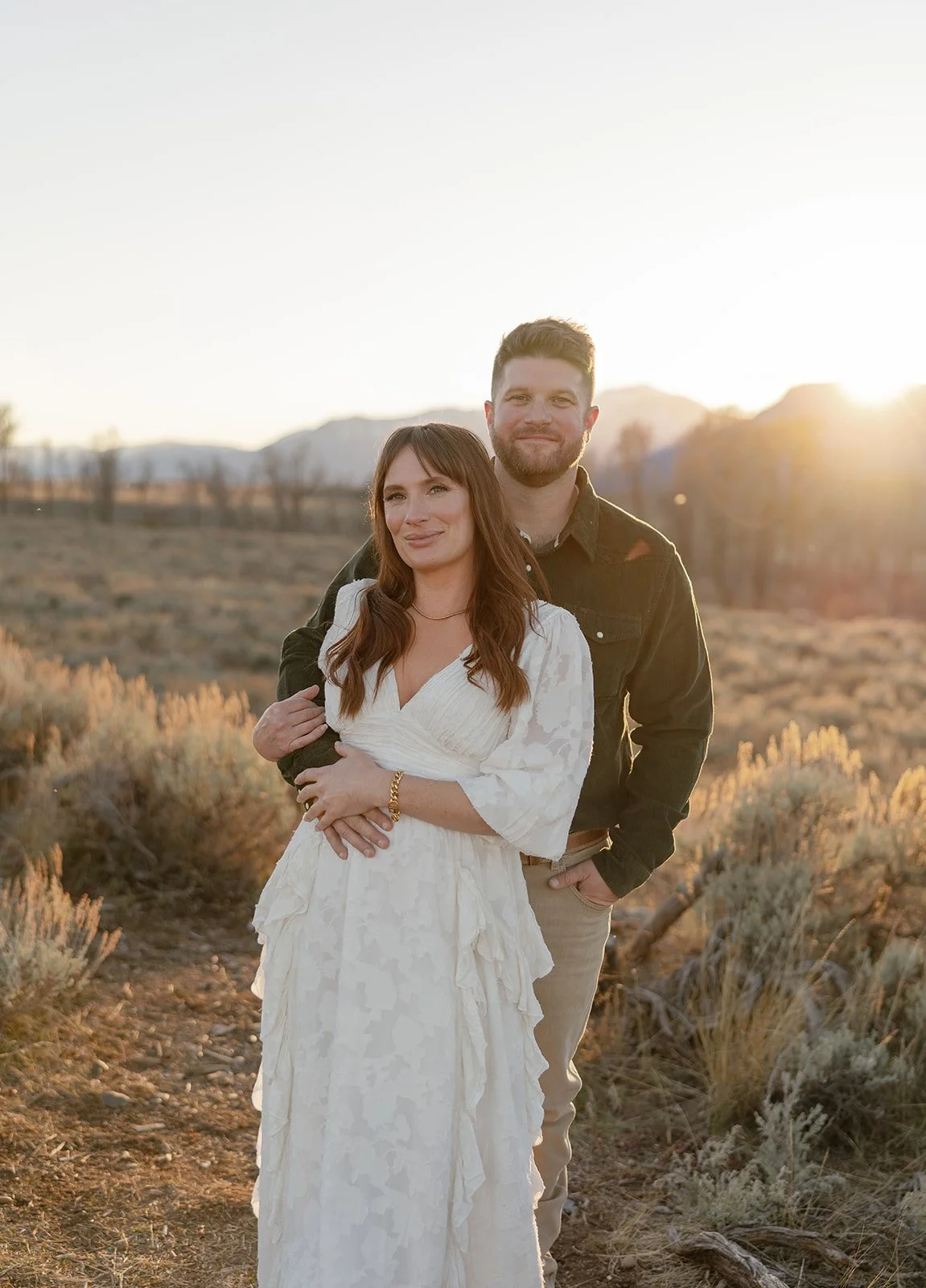 A couple standing outdoors at sunset in a desert landscape, with mountains in the background. The woman is wearing a white dress and the man is in a dark jacket and beige pants. They are smiling and embracing each other.