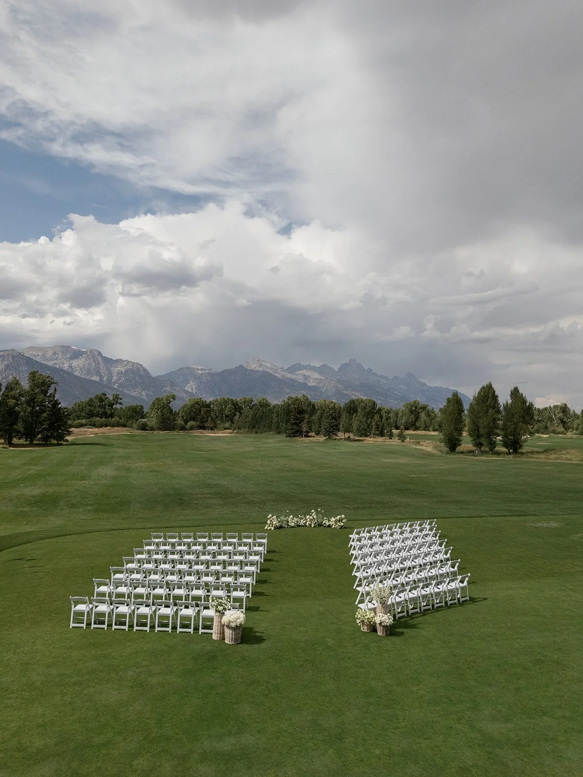 Wedding ceremony setup on a green outdoor field with white chairs arranged in two sections with floral decorations, set against a backdrop of mountains and a cloudy sky.