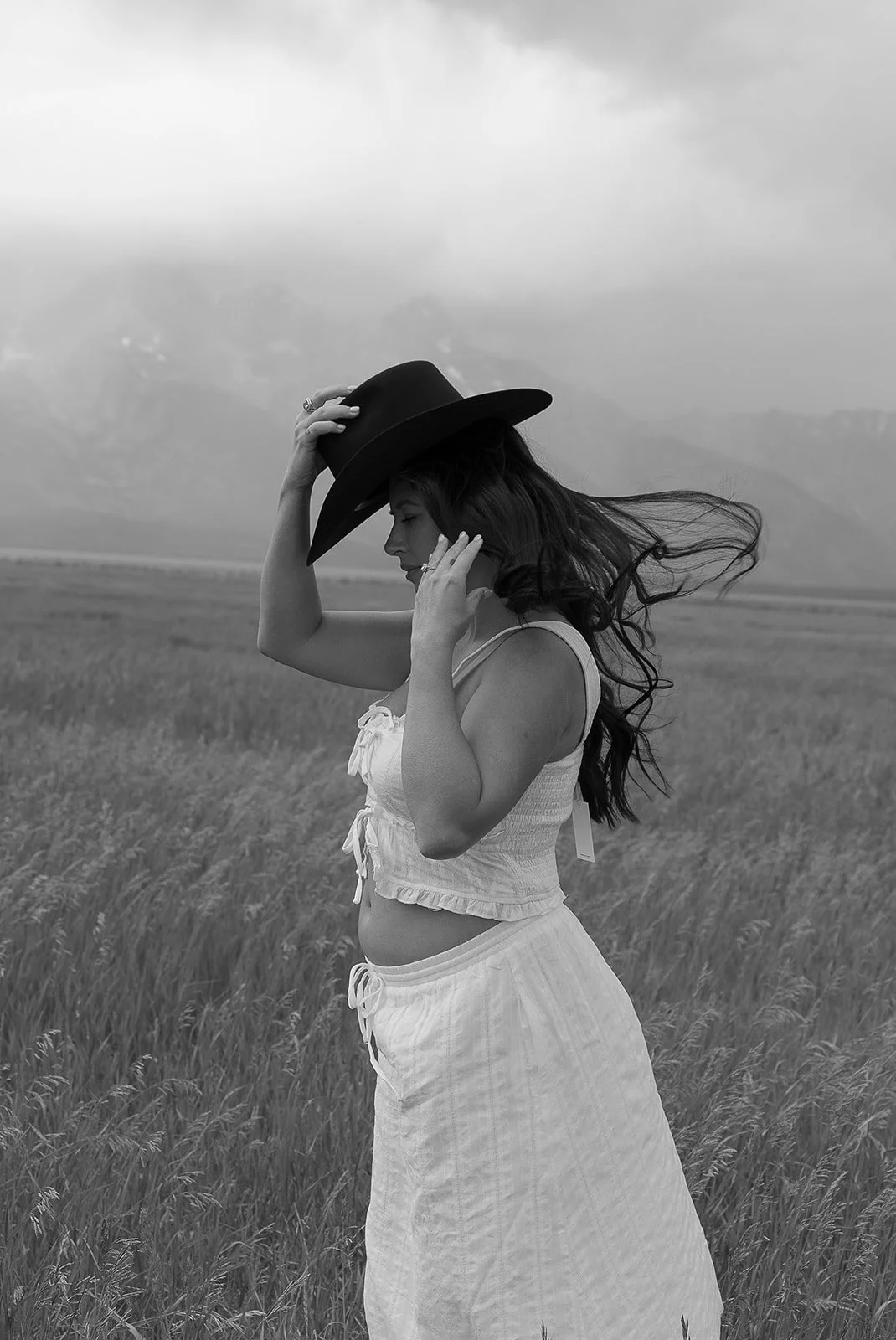A woman with long hair wearing a light-colored crop top and skirt, holding a wide-brimmed hat, standing in a grassy field with mountains in the background.