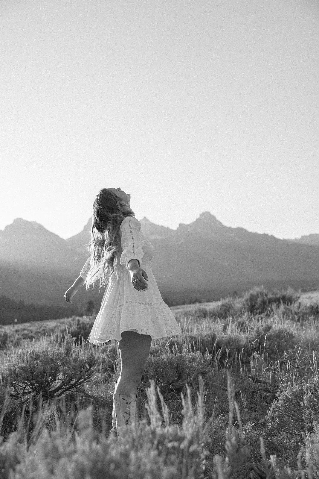 A woman in a white dress standing in a grassy field with mountains in the background, looking upward with arms outstretched.