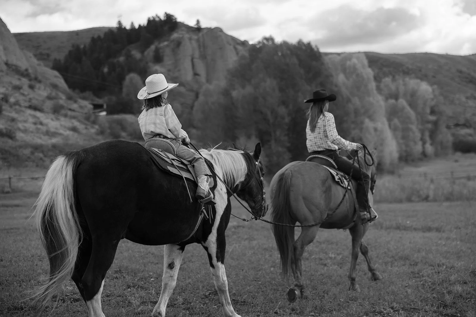 Two women riding horses across a grassy field with trees and cliffs in the background, wearing hats and casual clothing.