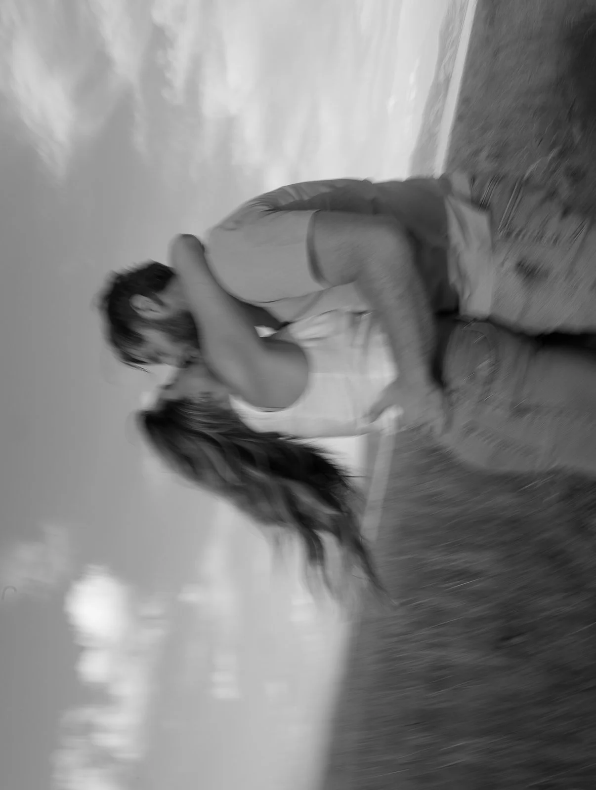 Two young women with long hair embracing outdoors on grassy terrain, with cloudy sky in the background.