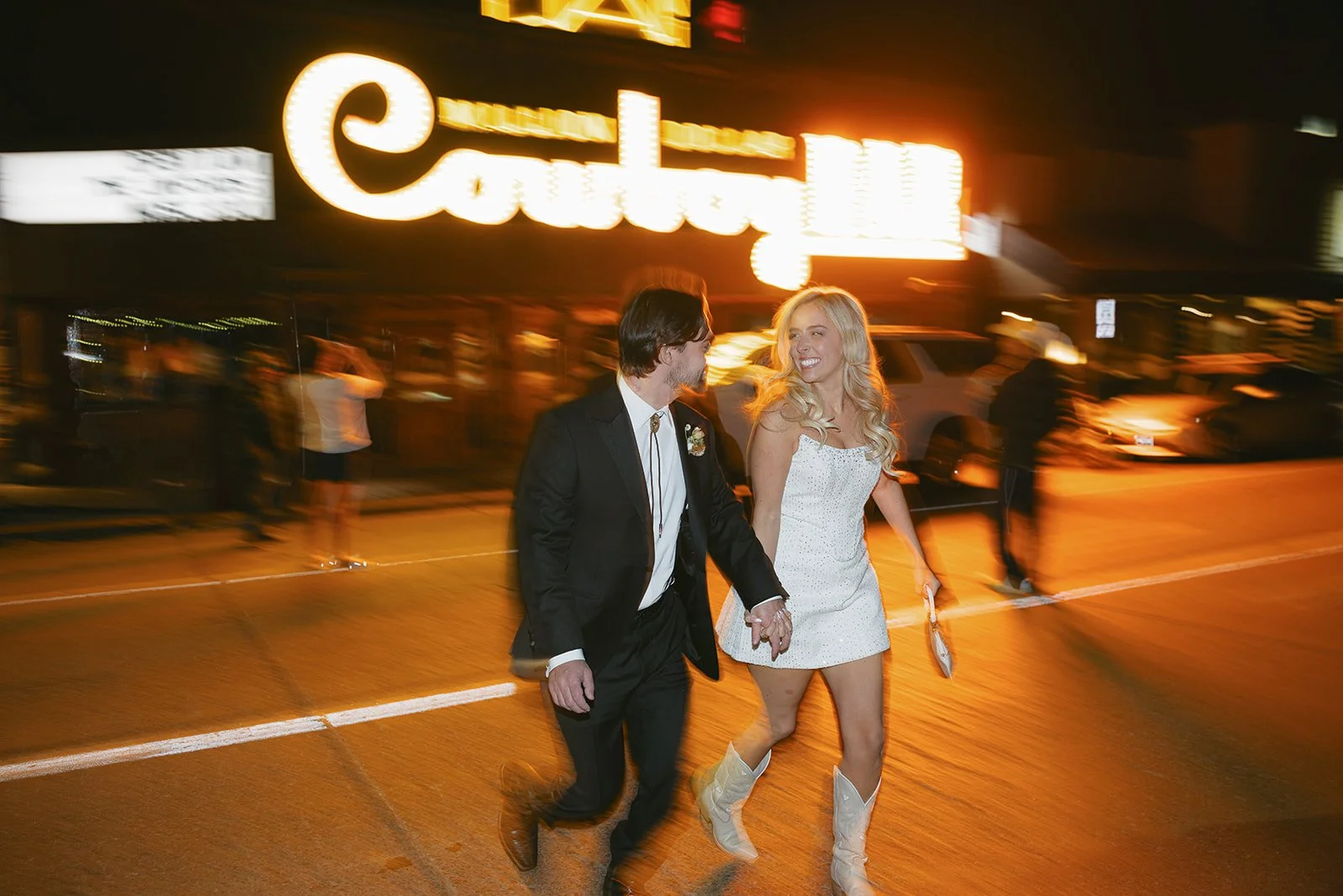 A couple dressed in formal attire holding hands and walking on a city street at night, with a brightly lit storefront and blurred figures in the background.