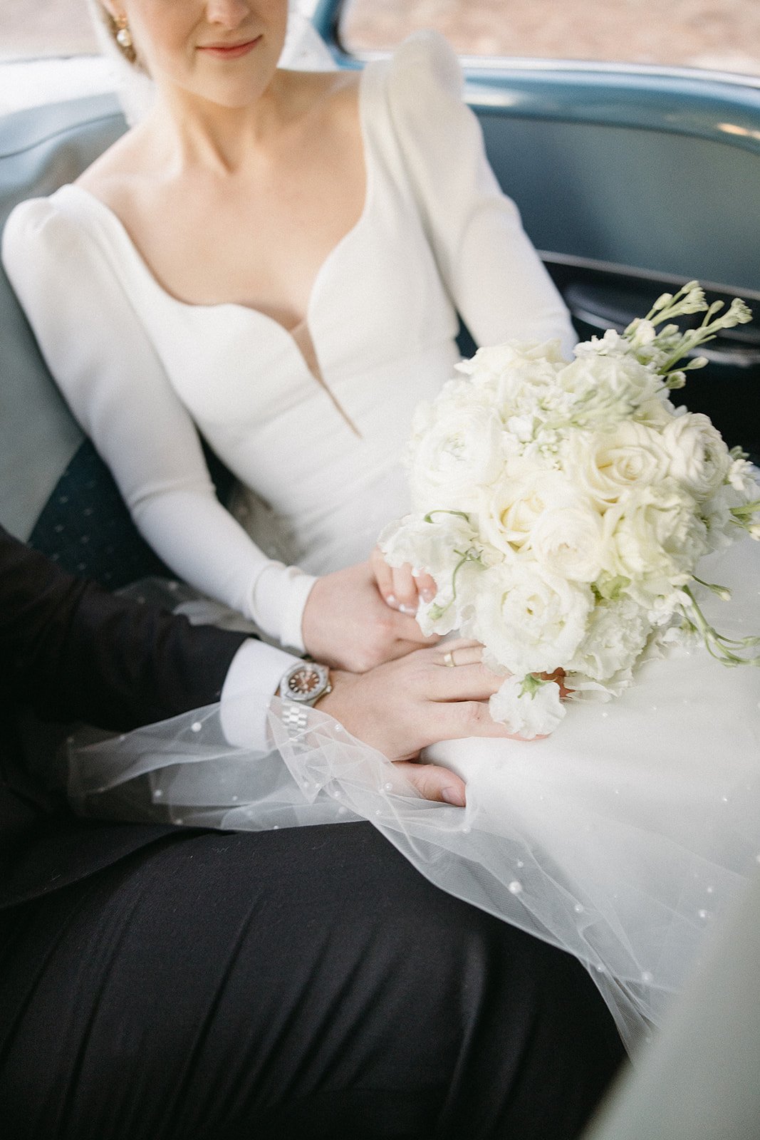 Bride in a white dress holding a bouquet of white flowers, sitting next to a person in a suit with hands lightly touching.