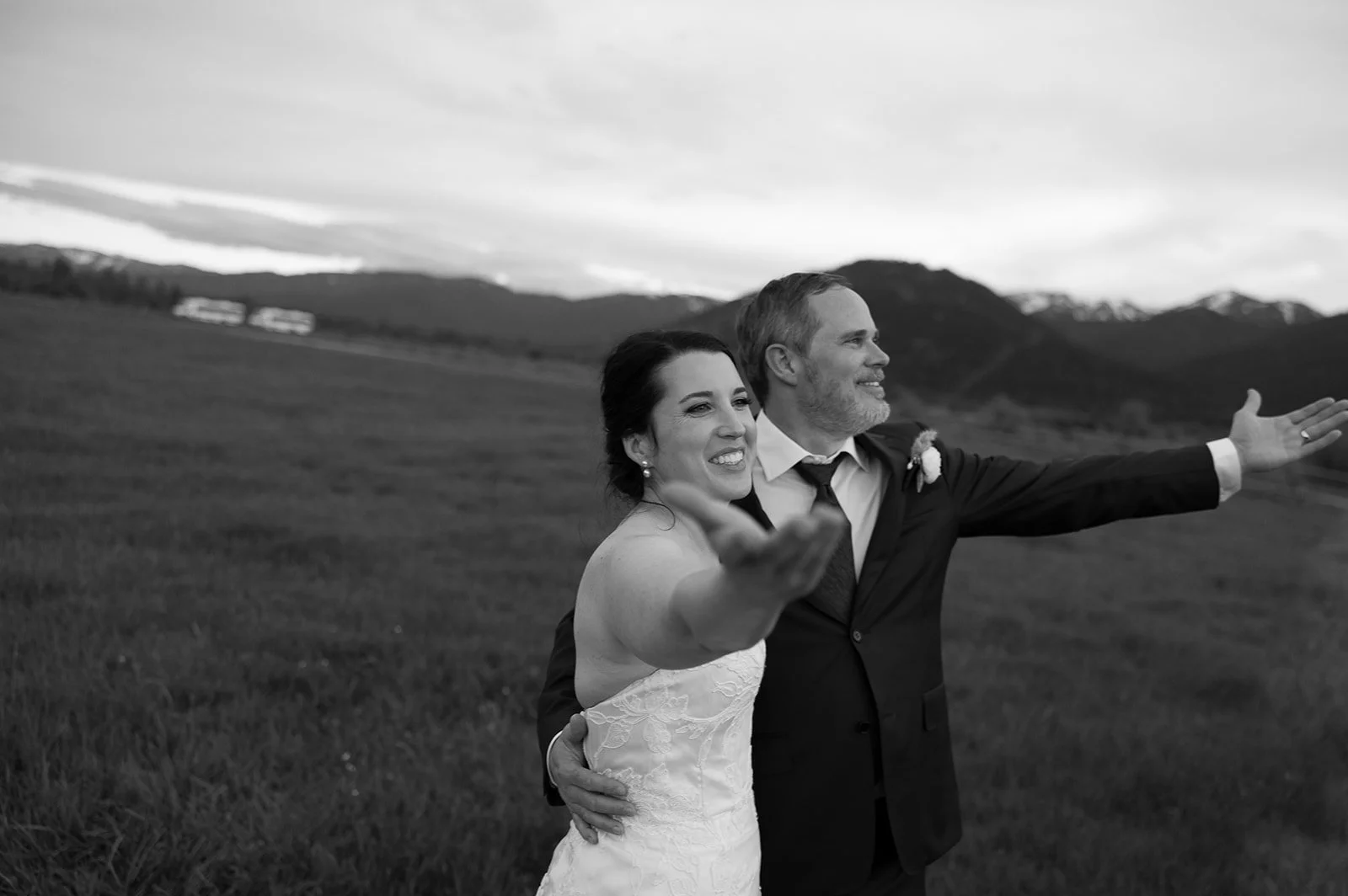 Black and white photo of a newlywed couple embracing outdoors with mountains in the background, both smiling and extending their arms.