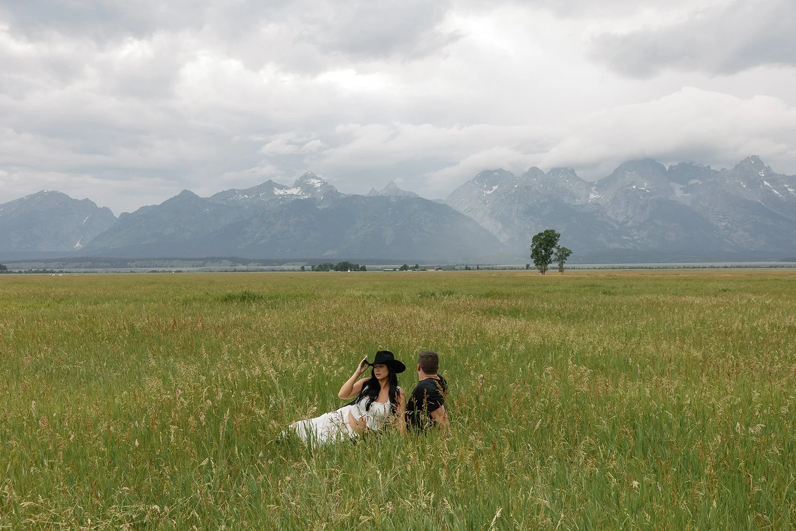 A woman and a man sitting in a green field with mountains in the background under cloudy skies. The woman is wearing a black hat and a white dress, while the man is wearing a dark shirt.