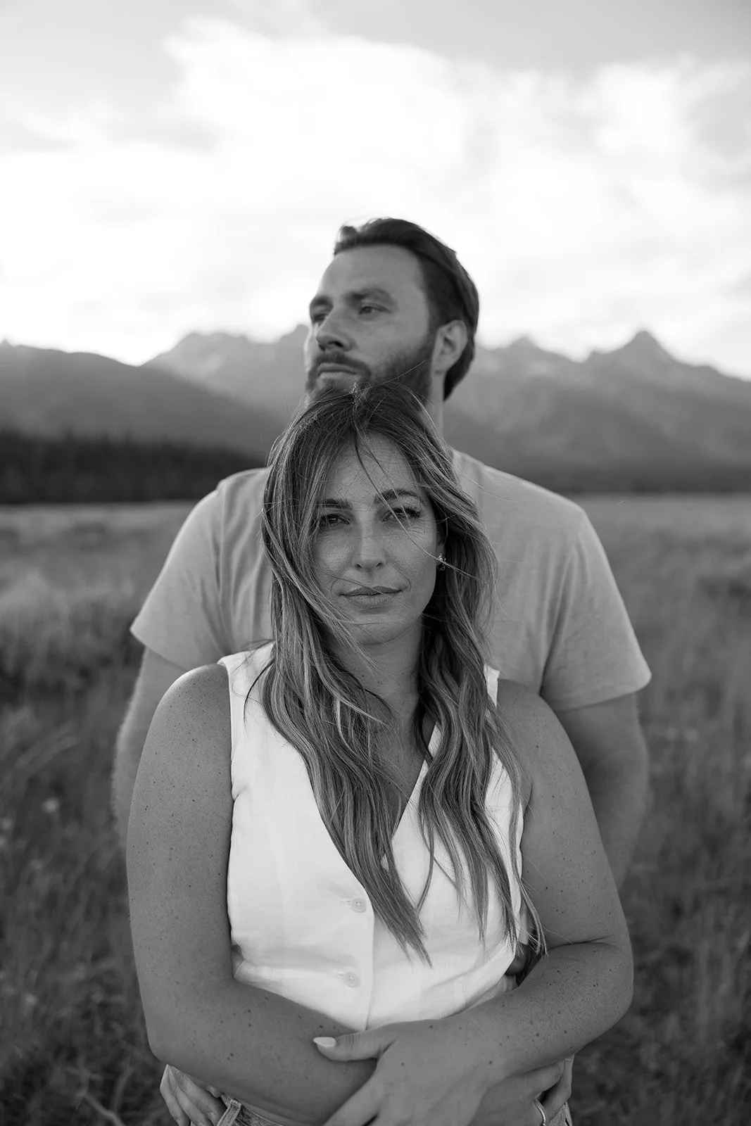 A black and white photo of a man and woman outdoors, with mountains in the background. The woman is in the foreground, with long wavy hair, wearing a sleeveless white top. The man is behind her, with a beard and slicked-back hair, wearing a t-shirt.