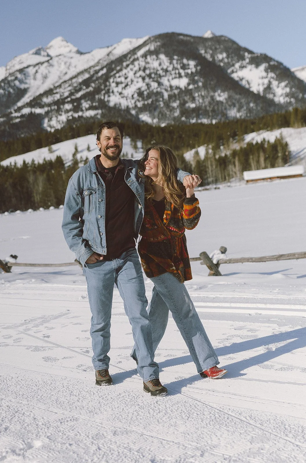 A man and woman smiling and walking in the snow with snow-capped mountains and trees in the background.