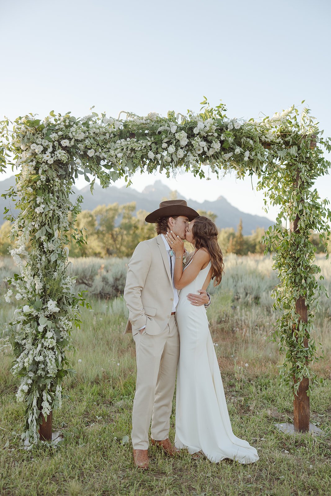 A couple dressed in wedding attire sharing a kiss under a floral arch outdoors with mountains in the background.