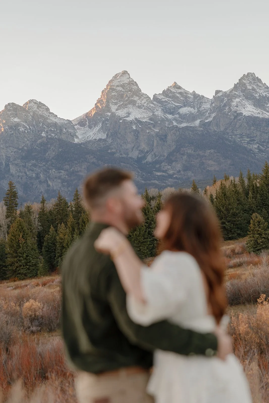 A blurry couple embracing outdoors with a mountain range and pine trees in the background.