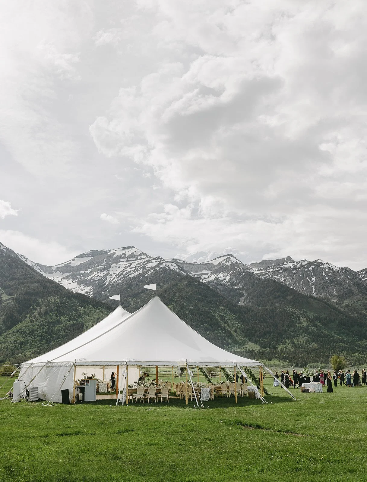 A large white event tent set up on a grassy field with outdoor tables and chairs, snow-capped mountains in the background, and a cloudy sky overhead.
