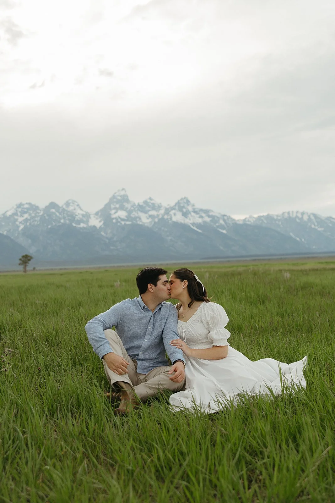 A couple sitting on grass in a field, kissing, with snow-capped mountains in the background.