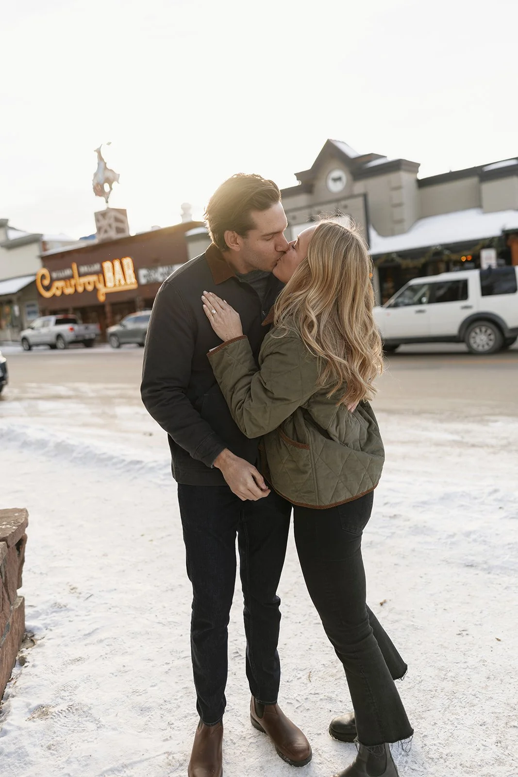 A couple sharing a kiss on a snowy street in front of a store with a sign that reads "Cabela's."