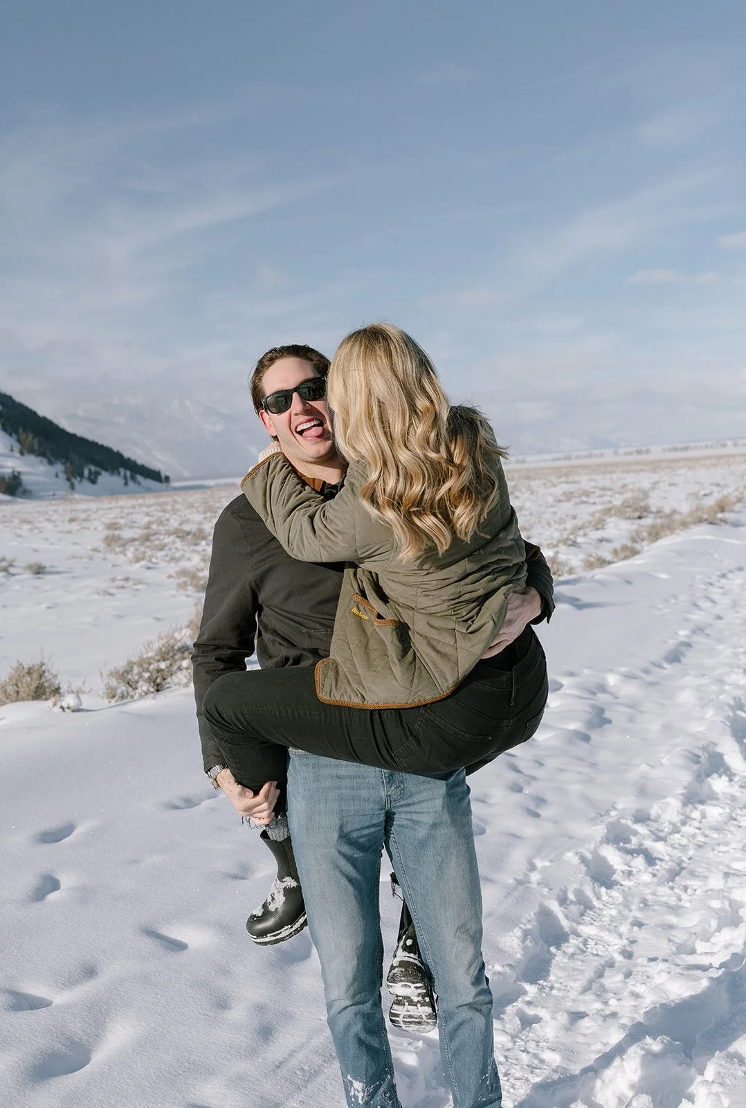 A man carrying a woman in a snowy landscape, both smiling and wearing winter clothing.