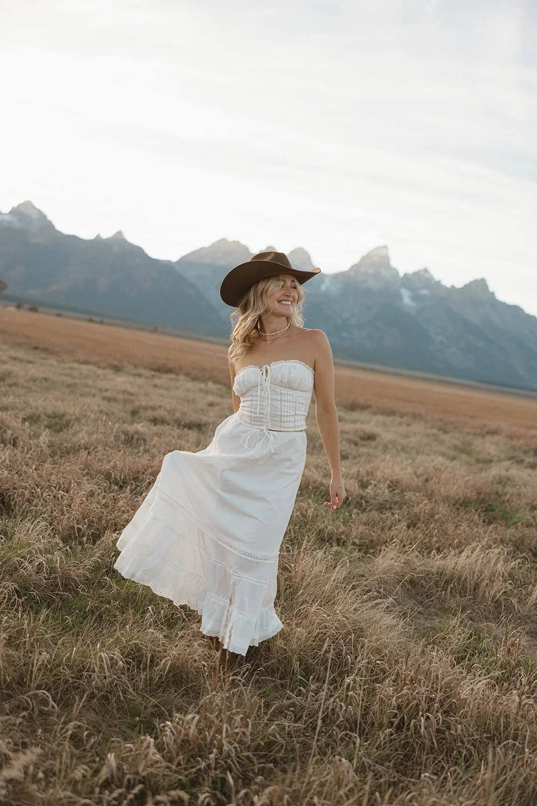 A woman in a white summer dress and a wide-brimmed brown hat standing in a field with mountains in the background.