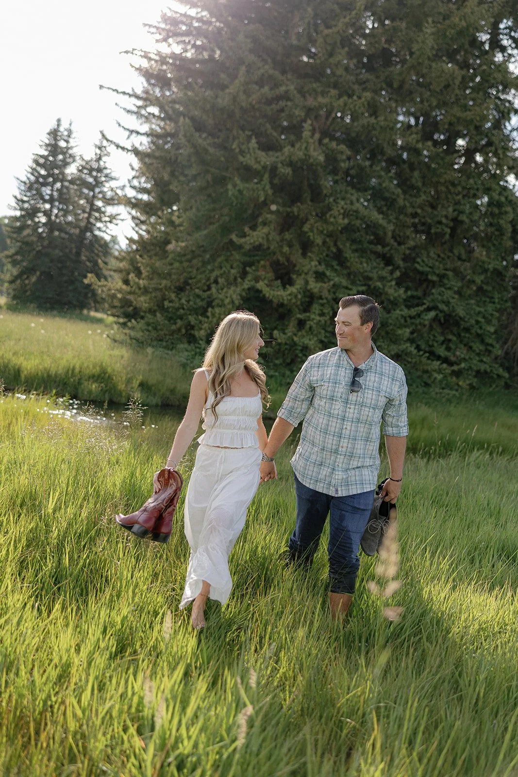 A couple walking hand in hand through a grassy field near a pond with tall trees in the background.