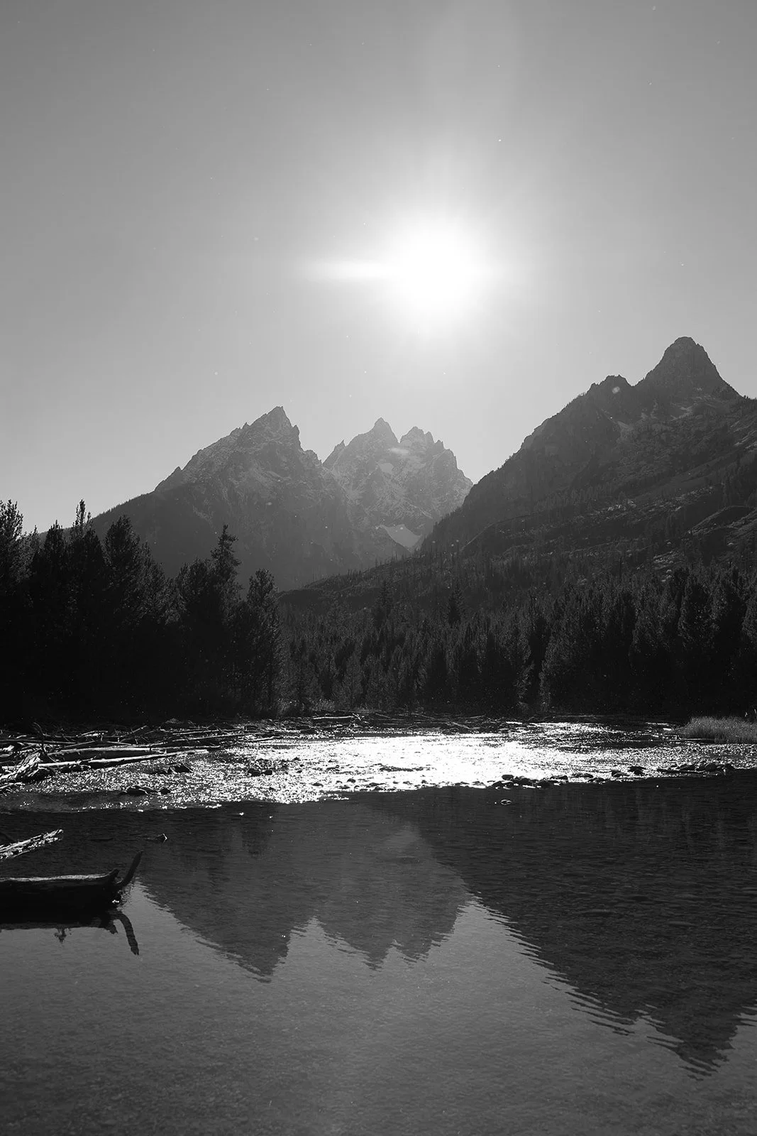 Black and white landscape photo of a mountain range with tall, jagged peaks, a river in the foreground reflecting the mountains and sky, trees along the riverbank, and the sun shining brightly overhead.