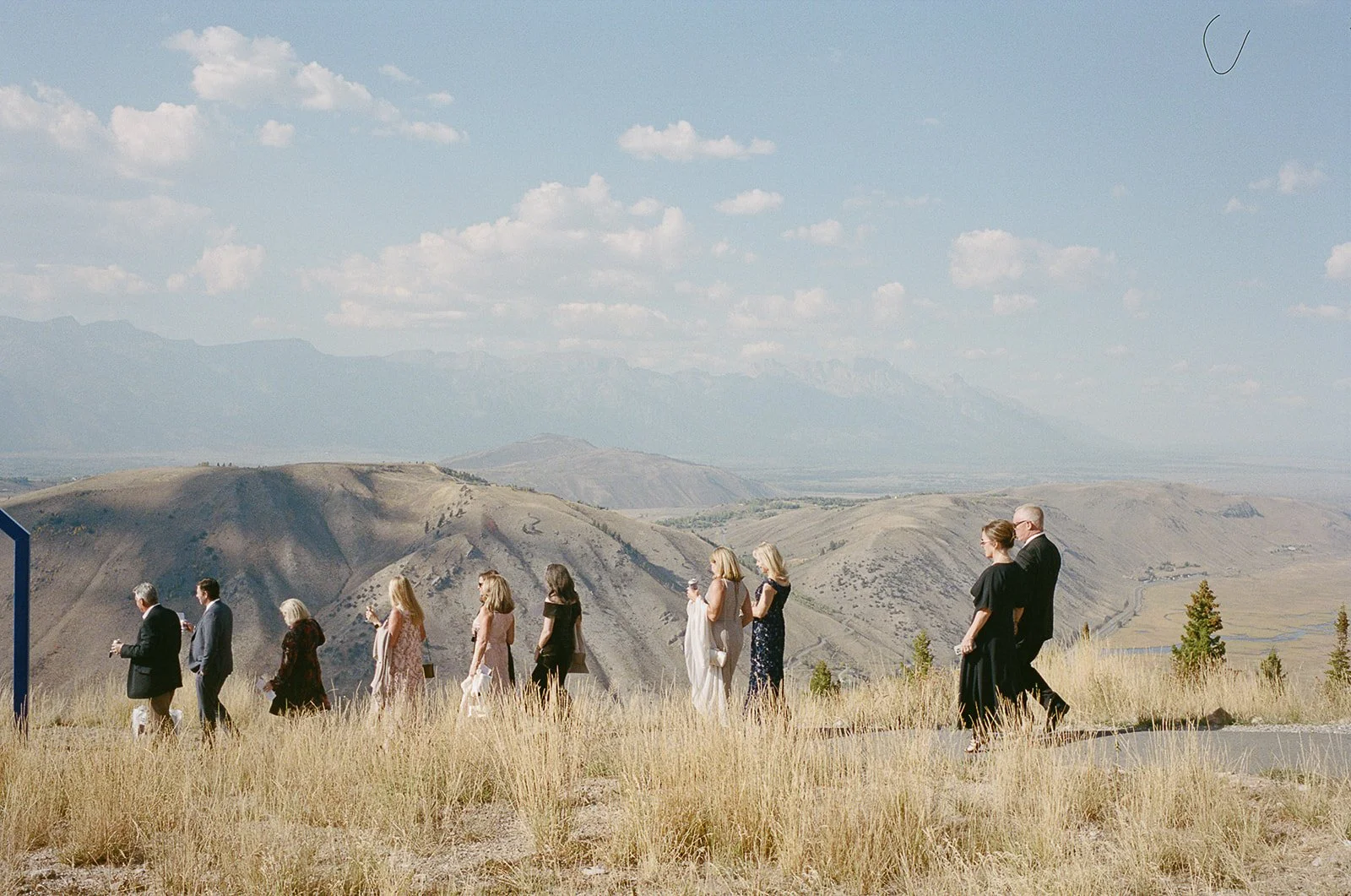 A group of people dressed in formal attire walking through a grassy, hilly landscape with mountains in the background.