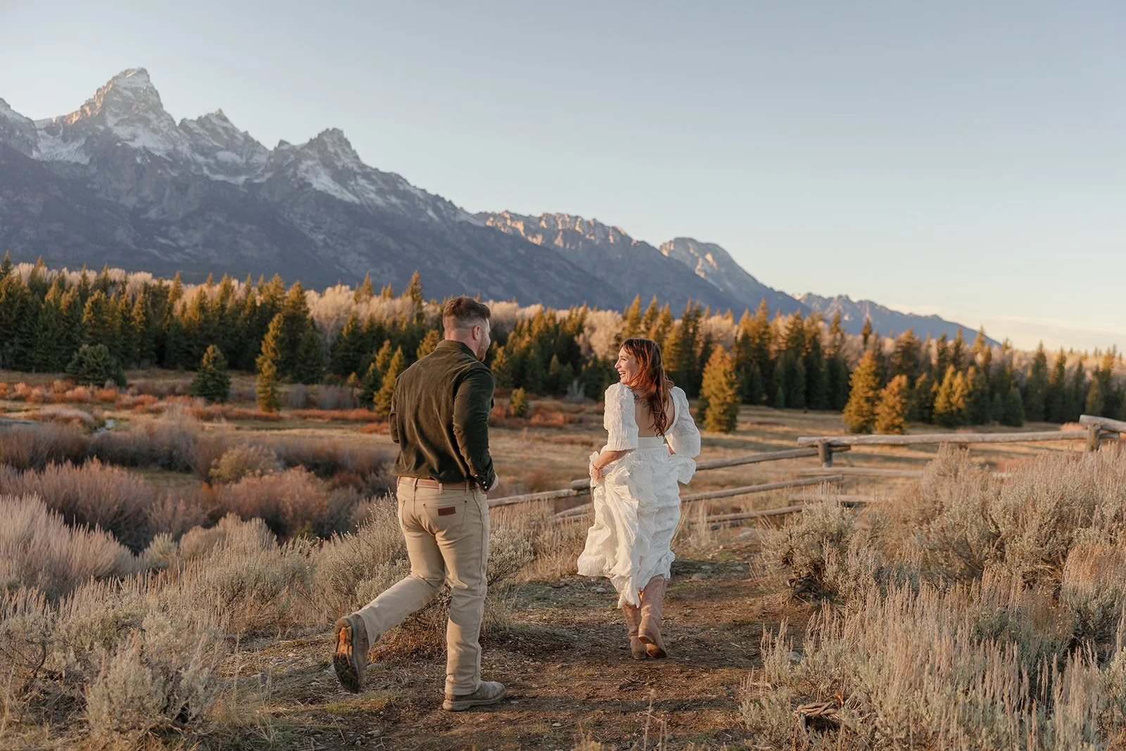 A couple walking along a dirt path in a mountain landscape at sunset. The woman is wearing a white dress, and the man is wearing a dark jacket and beige pants.