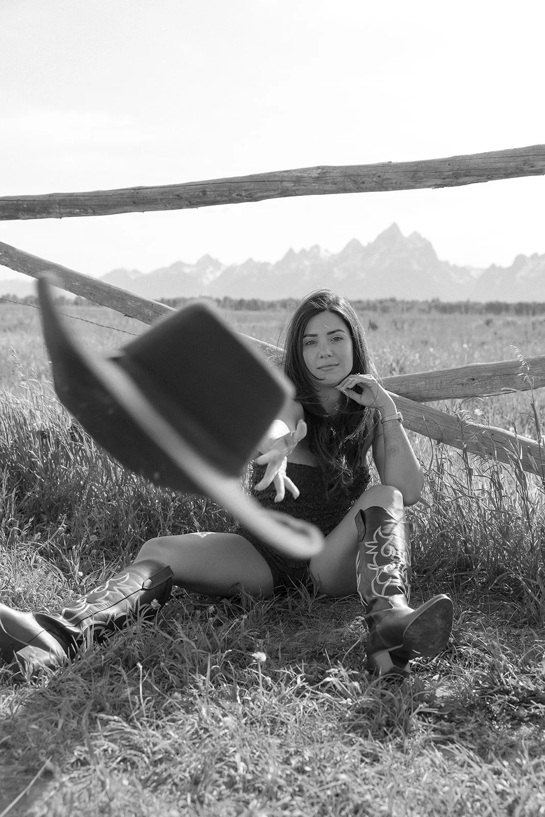 A woman sitting on grass in a field, reaching towards the camera with a hat in the foreground, wooden fence and mountains in the background, black and white photo.