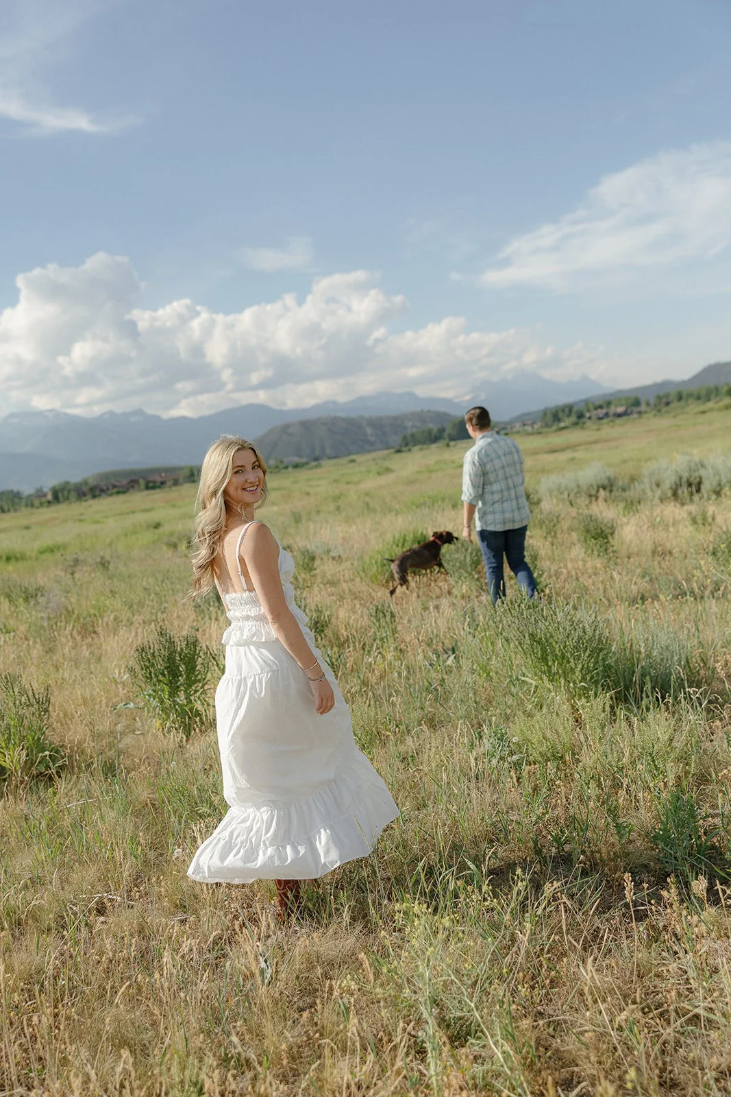 A woman in a white dress standing in a grassy field, with a man and dog in the background under a blue sky with clouds and mountains.