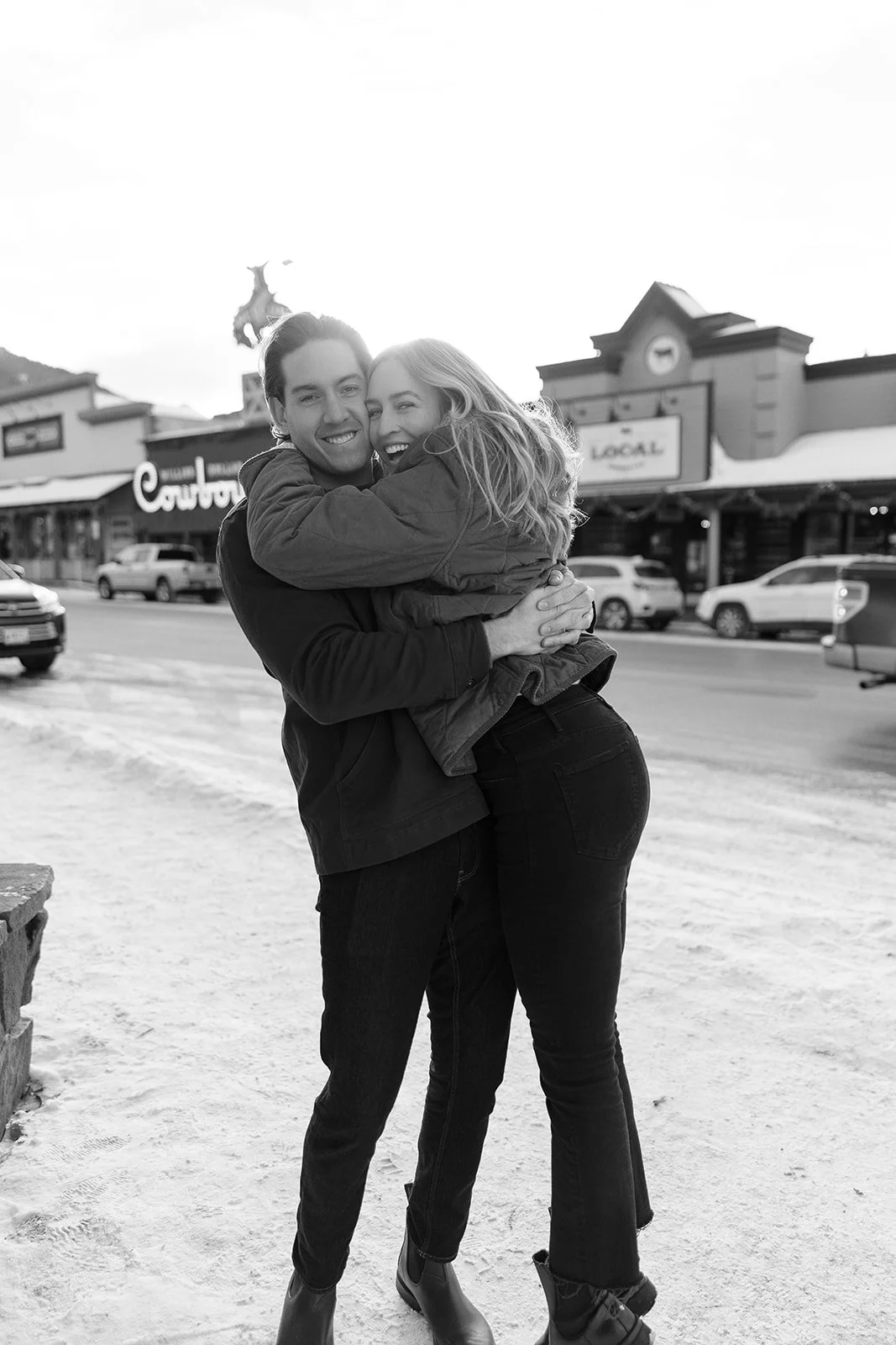 A black-and-white photo of a happy couple hugging outdoors on a snowy street.