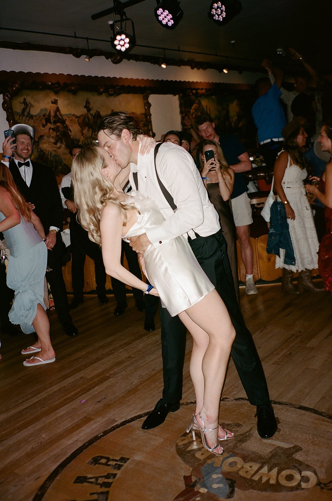 A romantic kiss between a man and a woman on the dance floor with party guests watching at a celebration or wedding reception.