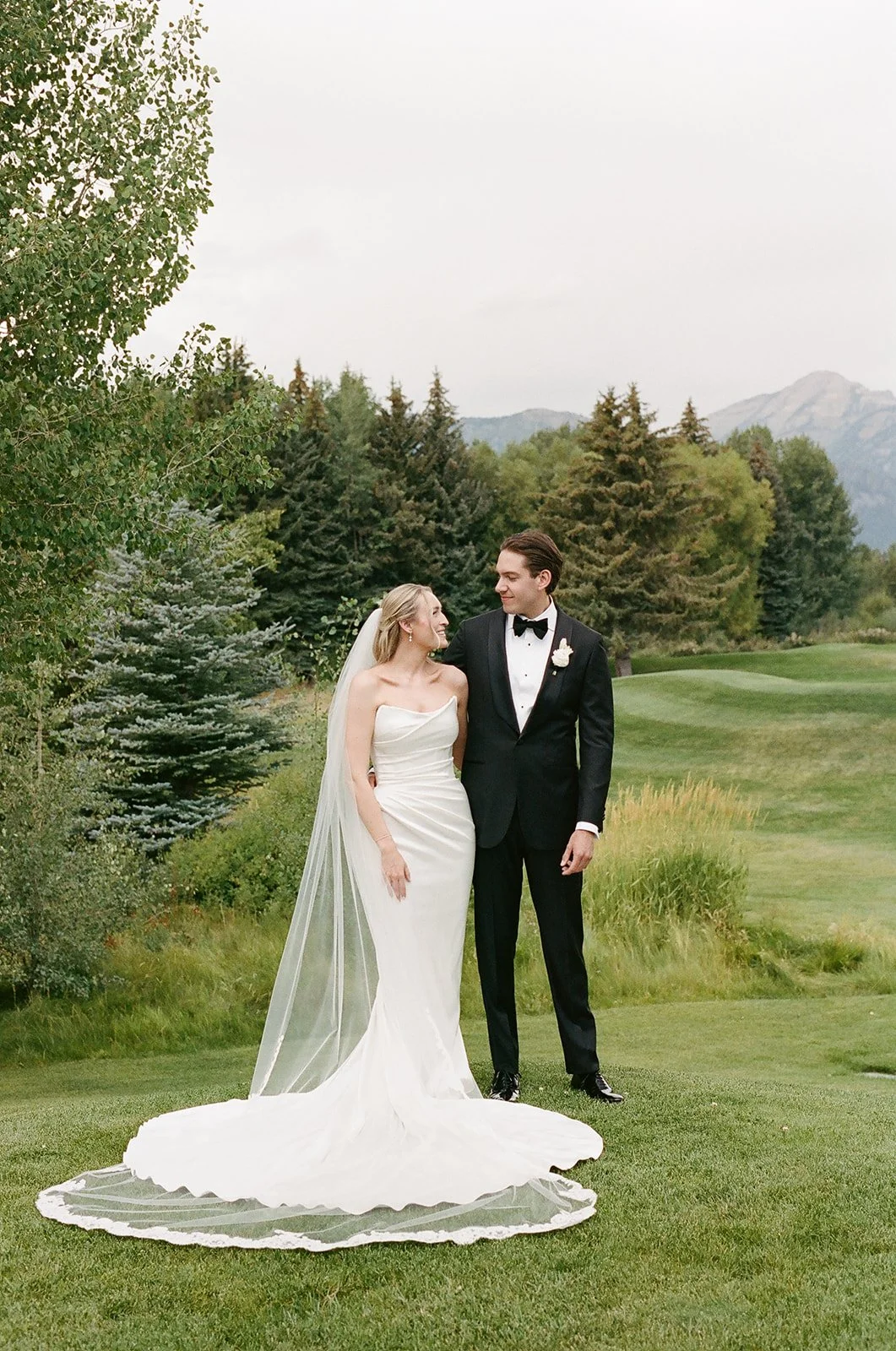 Bride and groom standing on a grassy area at a wedding, with trees and mountains in the background.