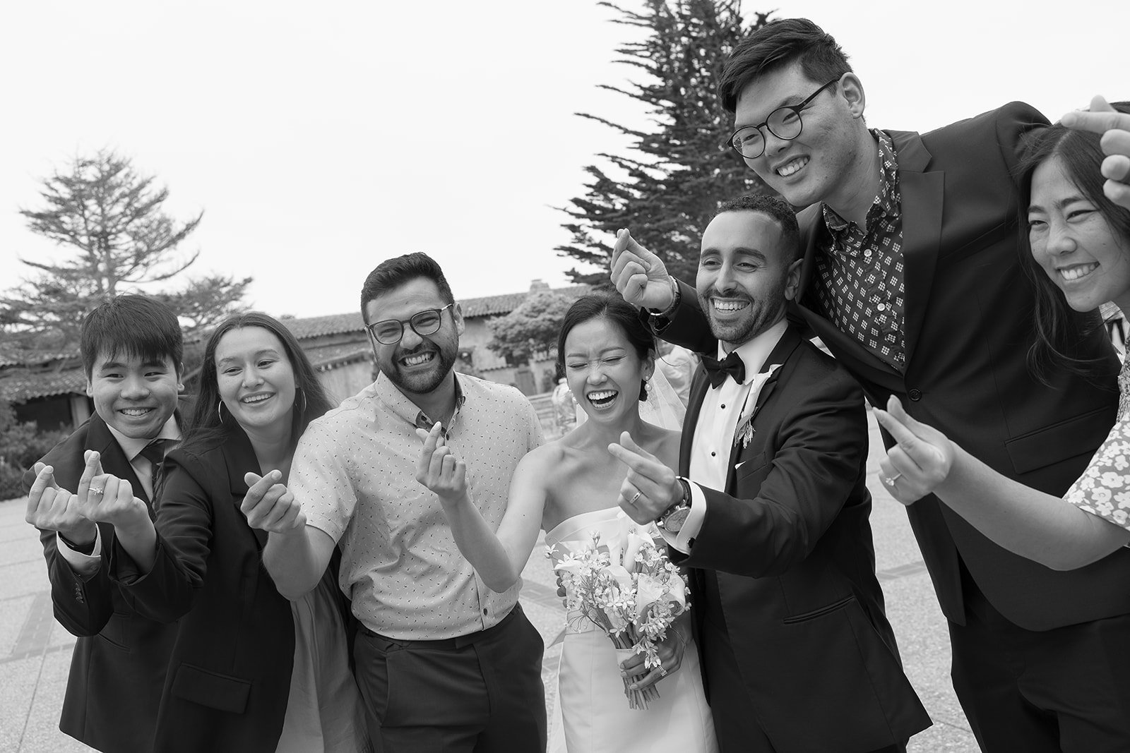 A group of six people, including a bride and groom, laughing and making finger heart gestures at an outdoor wedding.
