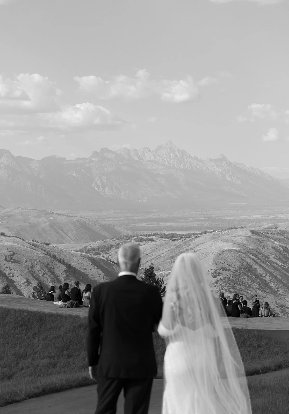 A black and white photo of a wedding ceremony outdoors with a mountain range in the background, a bride in a white wedding dress with a veil, and an older man in a dark suit walking towards the ceremony, with guests sitting on the grass.