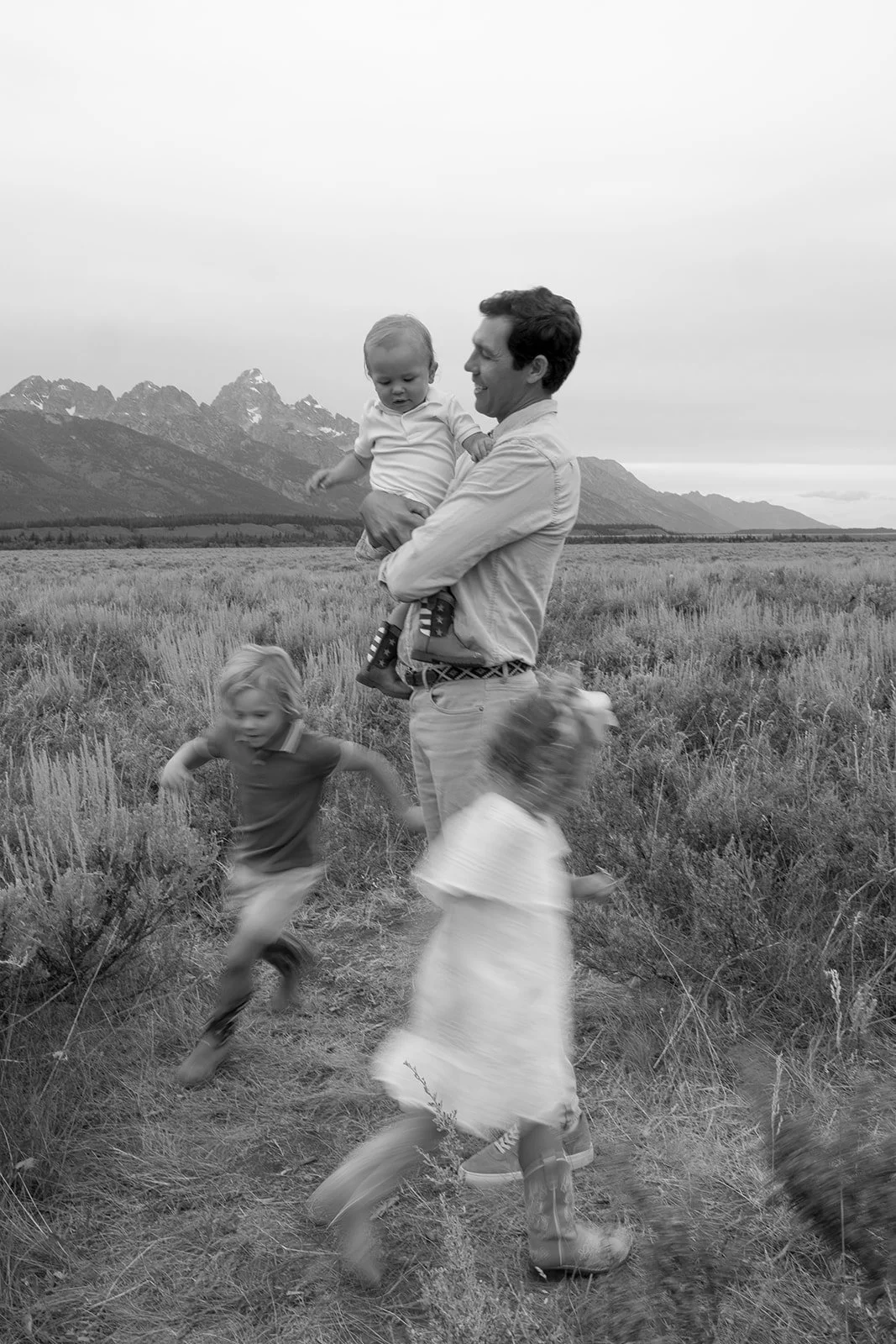 A man holding a young child in a field with mountain scenery in the background, while two other children run and play nearby.
