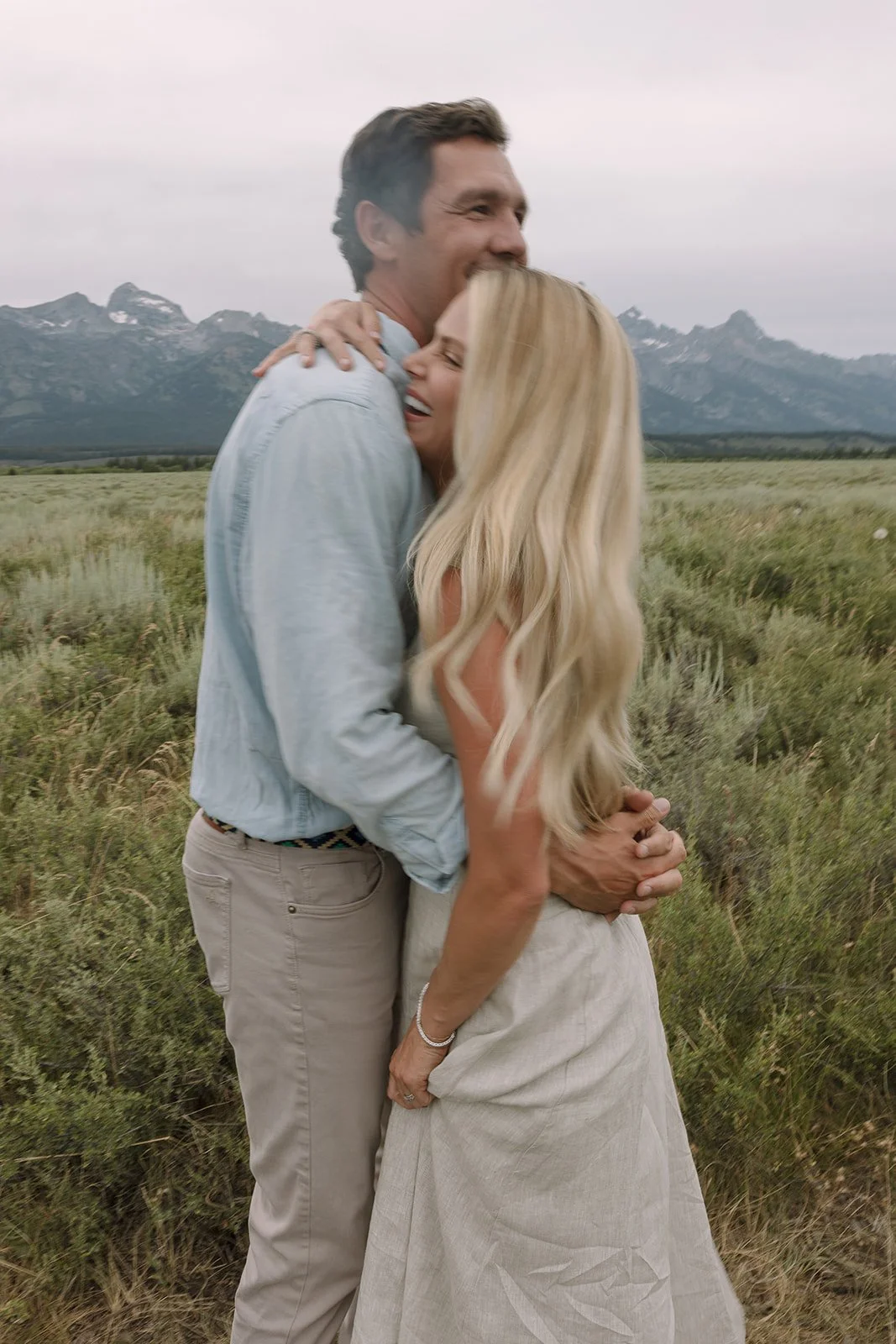 A man and woman embracing in a field with mountains in the background, both smiling and laughing.