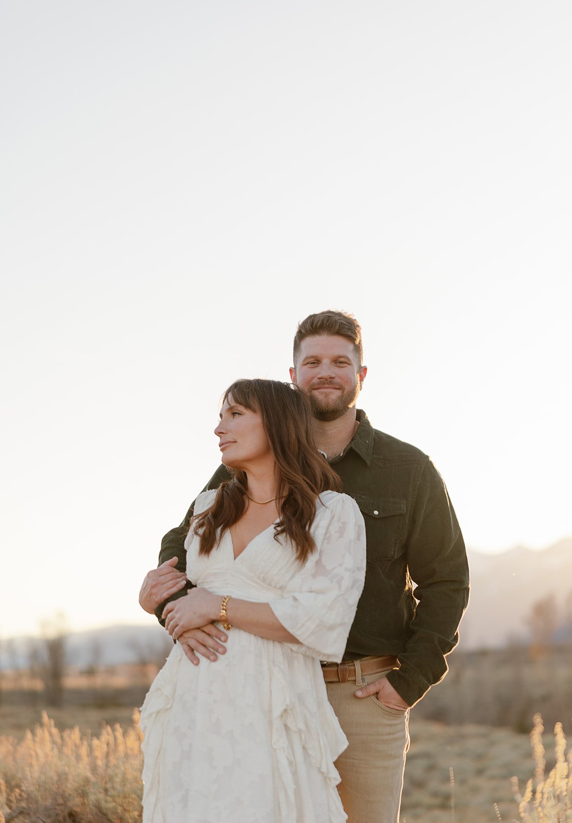 A couple standing outdoors at sunset, with the woman in a white dress and the man in a black shirt and tan pants, embracing each other.