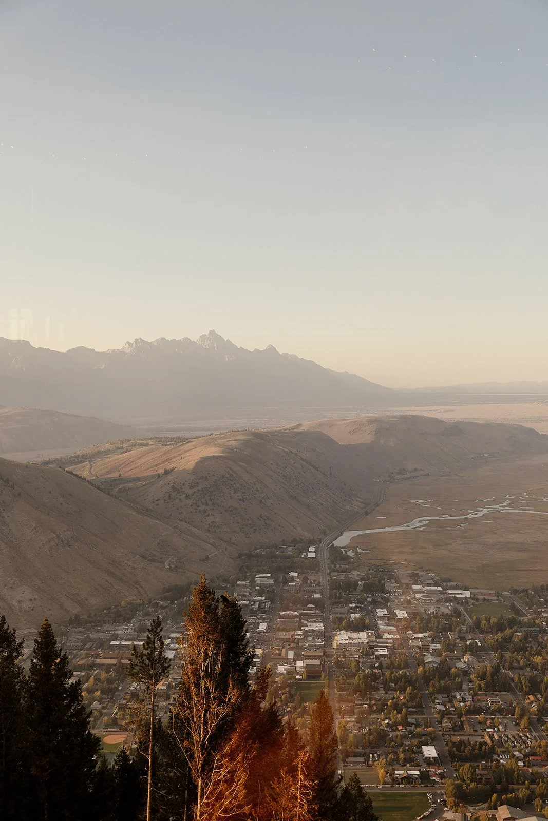 Aerial view of a town with houses, trees, and roads in a valley, surrounded by mountains under a clear sky.
