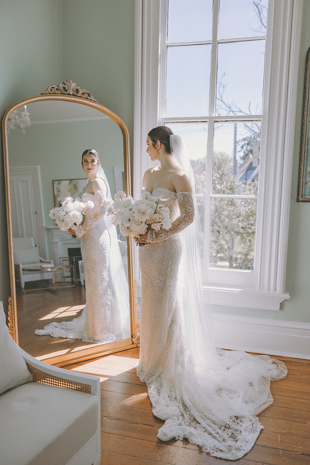 Bride in a lace wedding dress holding a bouquet, reflected in a large ornate mirror in a bright room.
