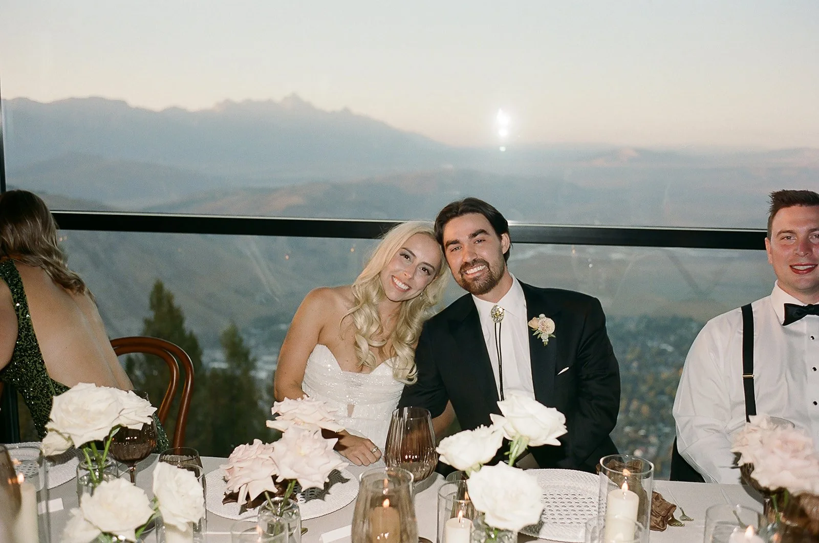 A wedding reception with a bride and groom sitting at a decorated table, smiling at the camera, with scenic mountains in the background.