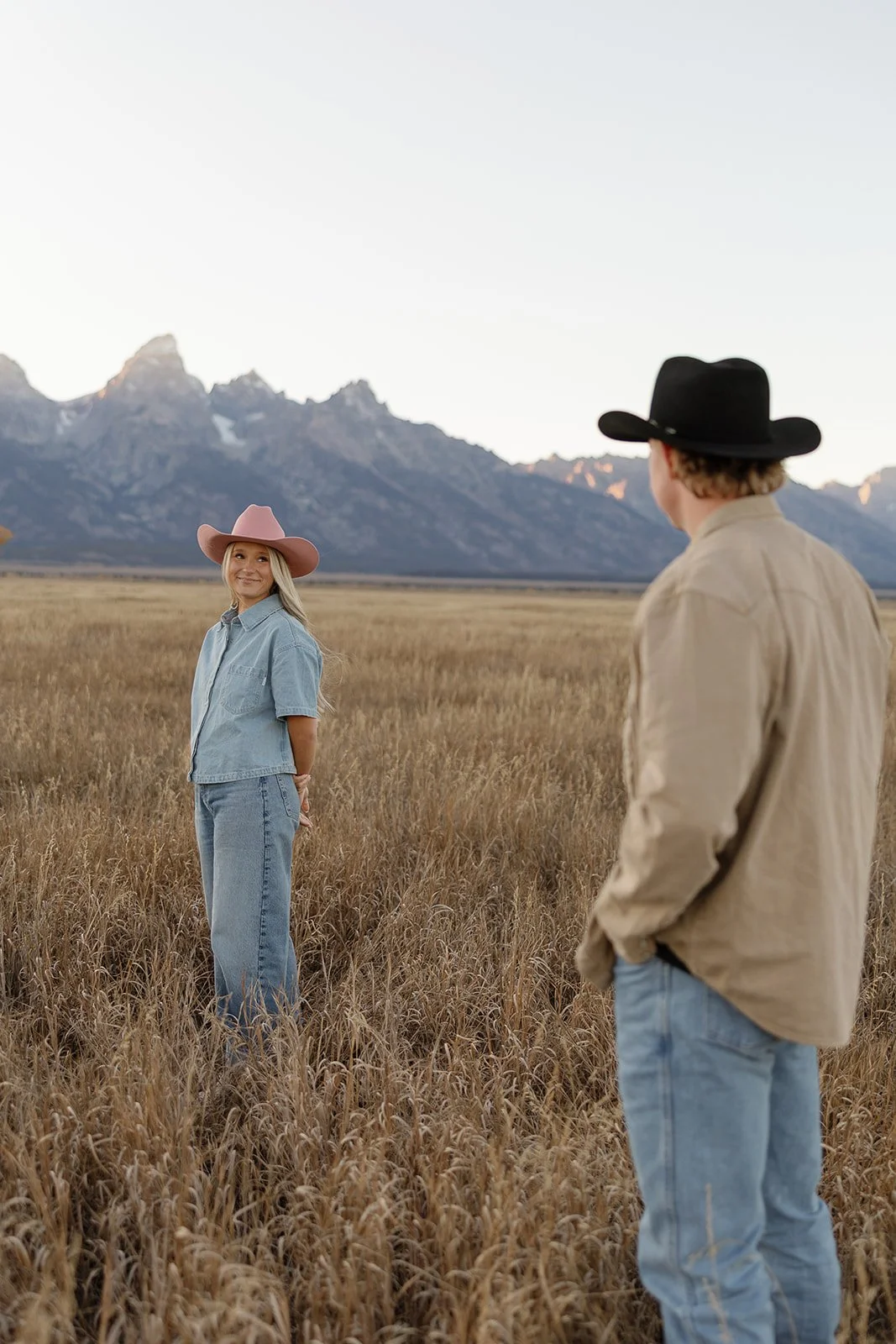 A woman standing in a field of tall grass, wearing a pink hat and denim outfit, smiling at a man in a black hat and beige shirt, with mountains in the background during sunset.
