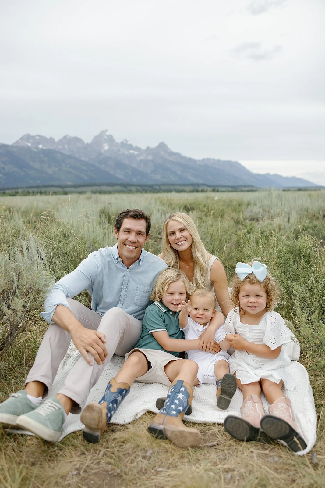 A family of six sitting on a blanket in a grassy field with mountains in the background. The family includes two adults and four children, all smiling at the camera.