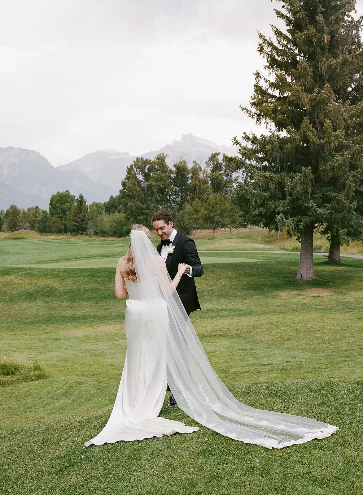 A bride and groom dancing on a grassy golf course with trees and mountains in the background, overcast sky.