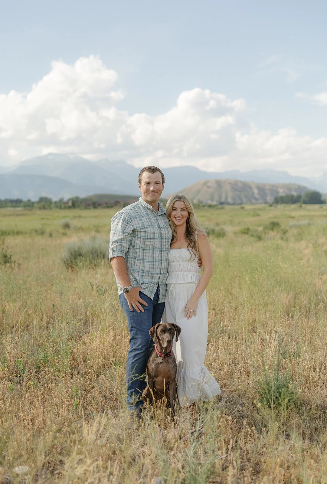 A couple standing in a grassy field with a dog, mountains and cloudy sky in the background.