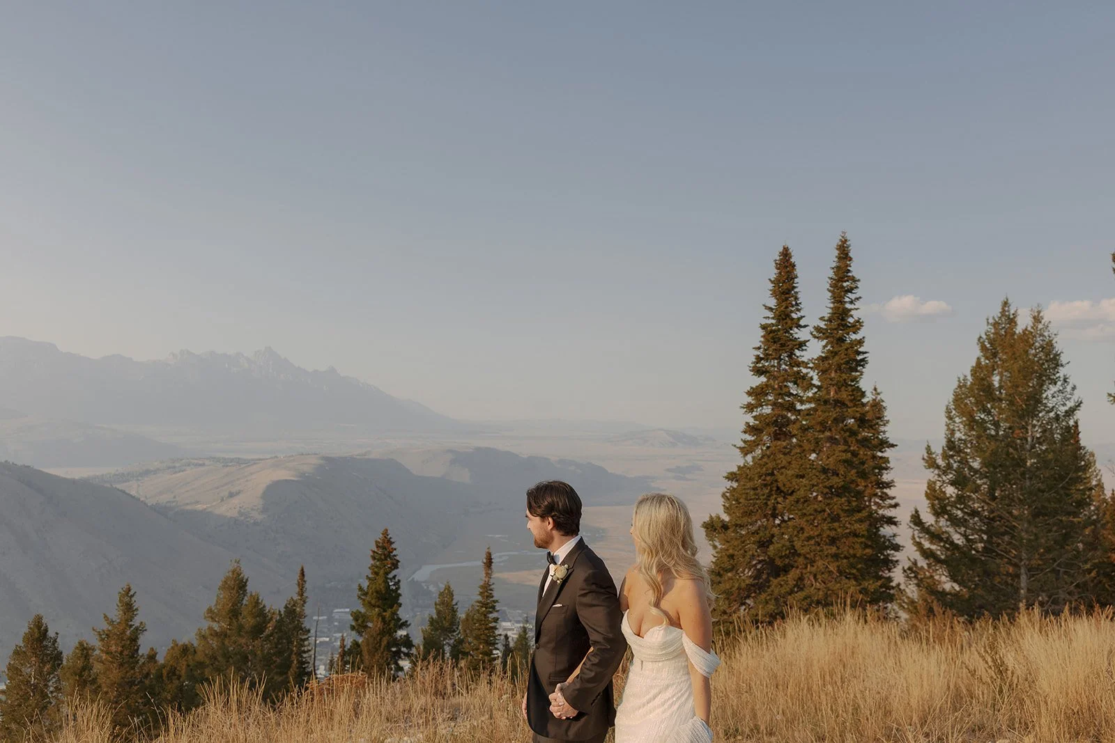 A bride and groom holding hands in a field of tall grass, with a mountain and forest landscape in the background during sunset.